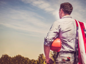 Attractive man in work clothes, holding tools and a US flag in his hands and looking into the distance against the background of trees, blue sky and sunset.