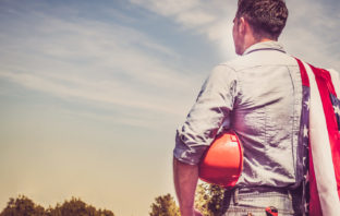 Attractive man in work clothes, holding tools and a US flag in his hands and looking into the distance against the background of trees, blue sky and sunset.